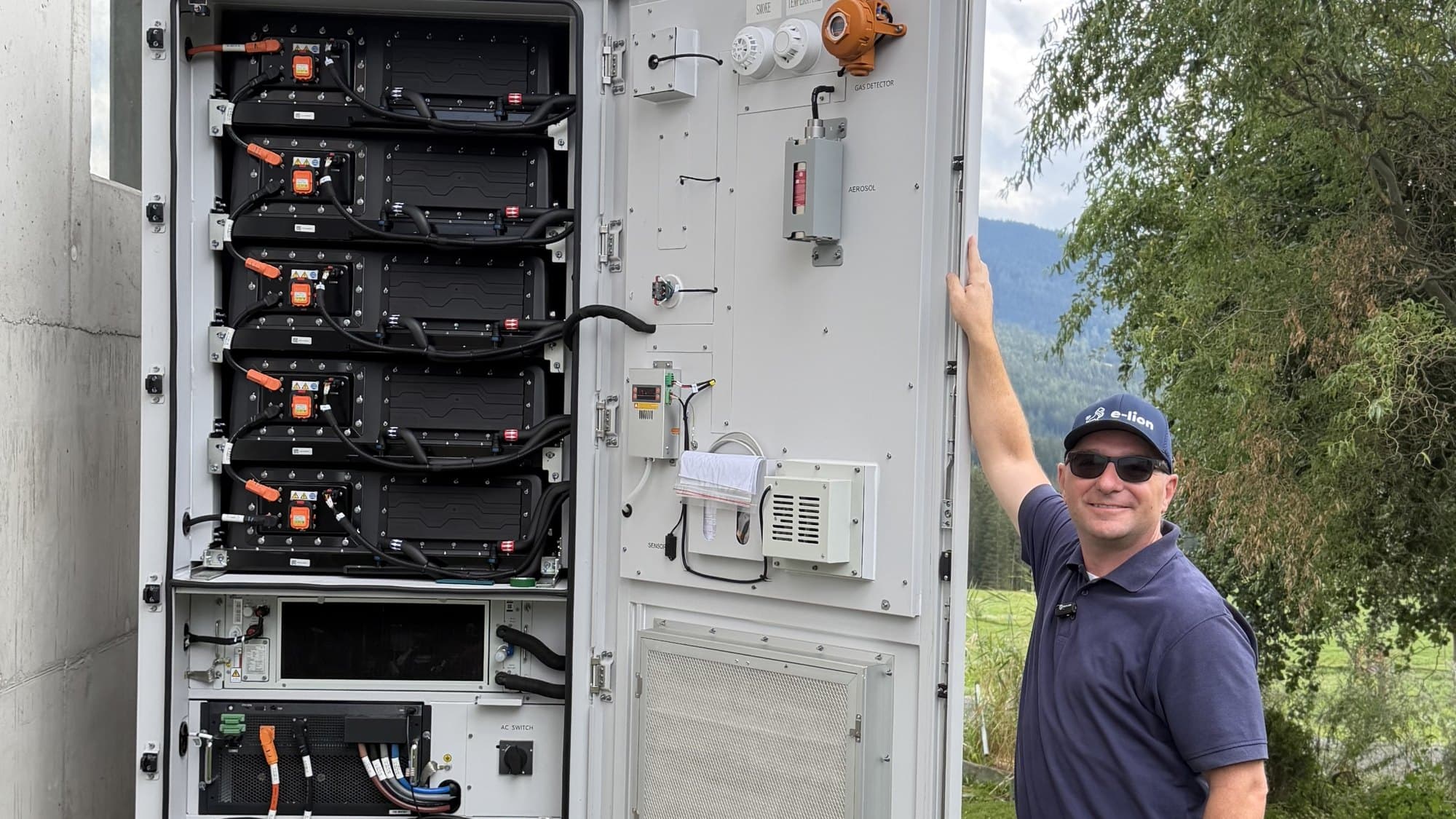 Geöffneter Risen-Gewerbespeicher mit Batteriemodulen, Gasmelder, Aerosol-Löschanlage und AC-Schalter, daneben ein e-lion-Mitarbeiter Geöffneter Risen-Gewerbespeicher mit Batteriemodulen, Gasmelder, Aerosol-Löschanlage und AC-Schalter, daneben ein e-lion-Mitarbeiter